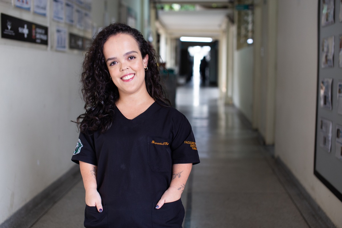 Na imagem, Fernanda, uma mulher branca com cabelos compridos e cacheados, tatuagens nos braços, usa uniforme preto. Ela está parada em um corredor de hospital, sorrindo para a  foto